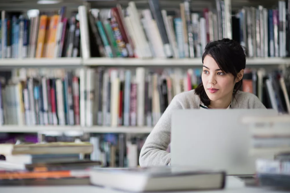 A woman in a library looks thoughtfully to the side, seated at a desk with an open laptop. Rows of colorful books fill the shelves behind her.